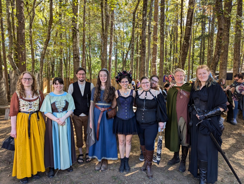 A group of people dressed in Renaissance festival attire stand together in a pretty, forested area.
